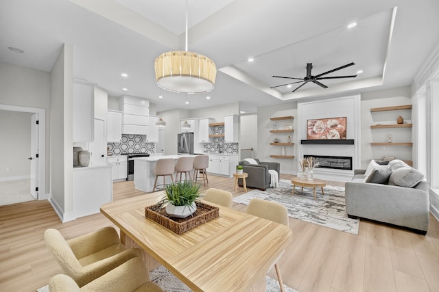 living room with ceiling fan, recessed lighting, a tray ceiling, light wood finished floors, and a glass covered fireplace