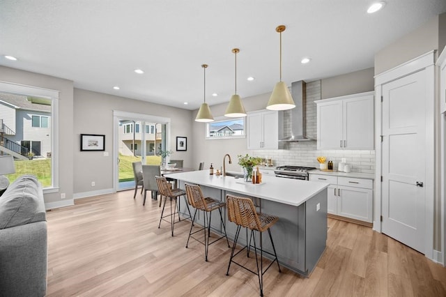 kitchen featuring white cabinetry, hanging light fixtures, an island with sink, wall chimney exhaust hood, and light hardwood / wood-style flooring