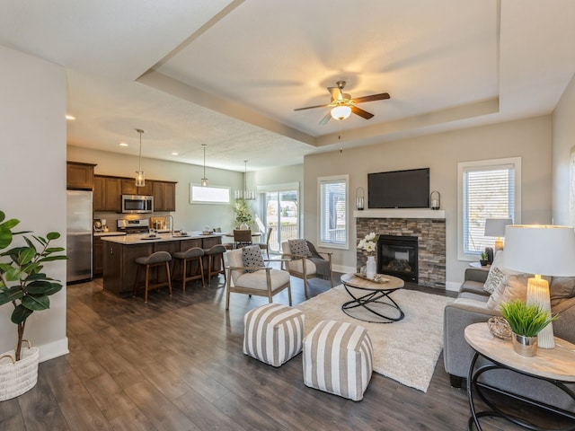 living room with a stone fireplace, dark hardwood / wood-style flooring, ceiling fan, and a tray ceiling