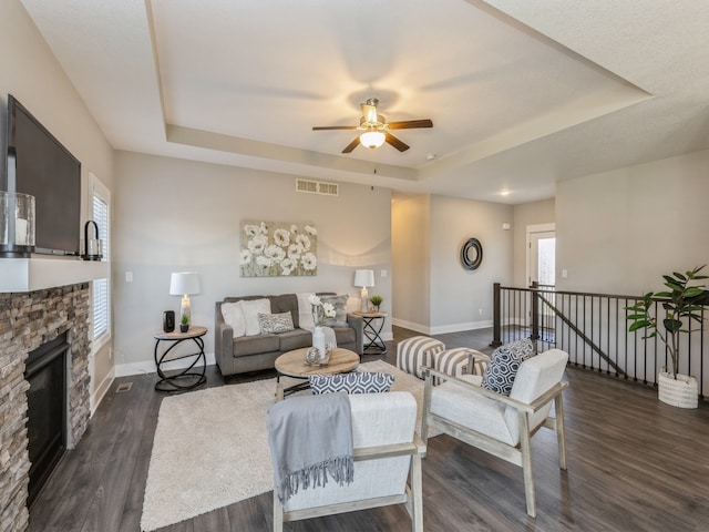 living room with a stone fireplace, ceiling fan, dark hardwood / wood-style floors, and a raised ceiling