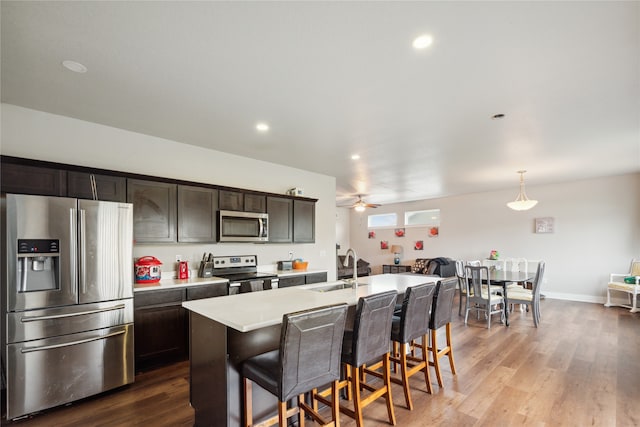 kitchen featuring stainless steel appliances, a center island with sink, hardwood / wood-style floors, hanging light fixtures, and ceiling fan