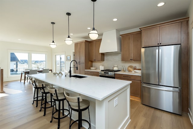 kitchen with stainless steel appliances, light wood-style floors, a sink, an island with sink, and premium range hood