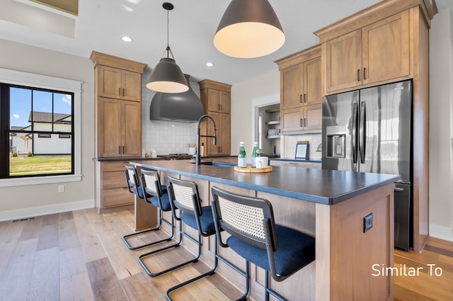 kitchen with hanging light fixtures, decorative backsplash, stainless steel fridge, an island with sink, and light hardwood / wood-style floors