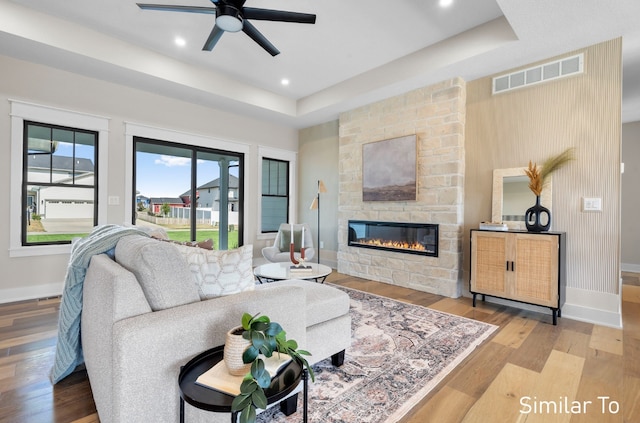 living room with ceiling fan, a stone fireplace, a tray ceiling, and light hardwood / wood-style flooring