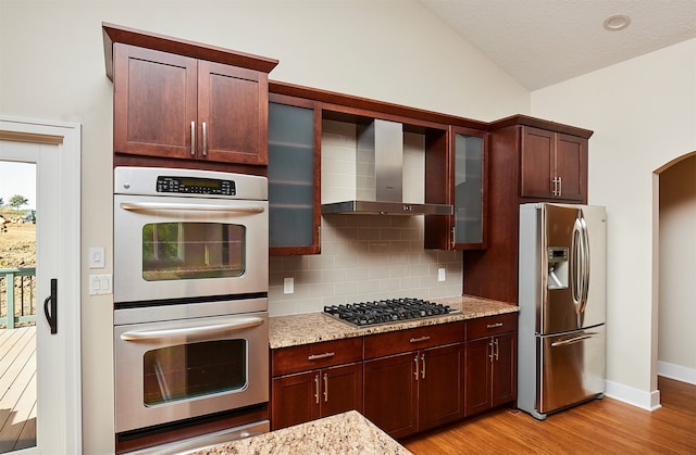 kitchen with light stone countertops, appliances with stainless steel finishes, light wood-type flooring, wall chimney exhaust hood, and vaulted ceiling