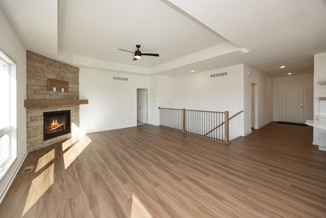 unfurnished living room featuring ceiling fan, a stone fireplace, a raised ceiling, and light wood-type flooring