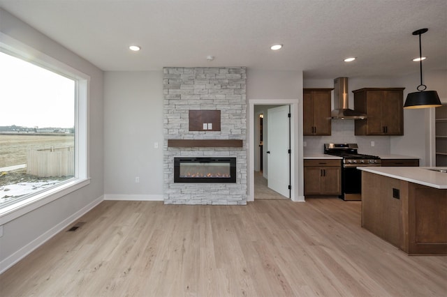 kitchen with a healthy amount of sunlight, hanging light fixtures, wall chimney range hood, stainless steel gas stove, and a stone fireplace