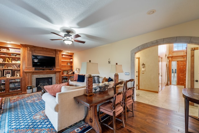 living room featuring built in shelves, a textured ceiling, ceiling fan, wood-type flooring, and a tiled fireplace