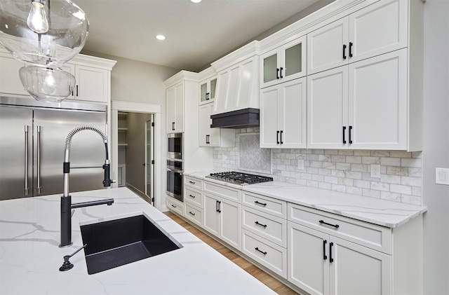 kitchen featuring white cabinets, a sink, backsplash, built in appliances, and glass insert cabinets