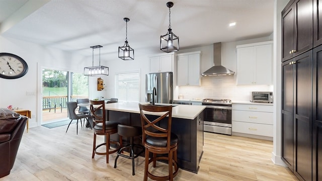 kitchen with stainless steel appliances, wall chimney range hood, decorative light fixtures, a center island, and light hardwood / wood-style floors