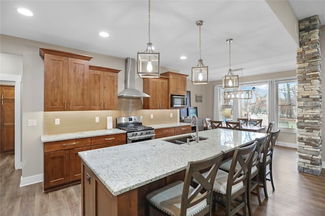 kitchen with sink, wall chimney exhaust hood, an island with sink, appliances with stainless steel finishes, and a kitchen bar