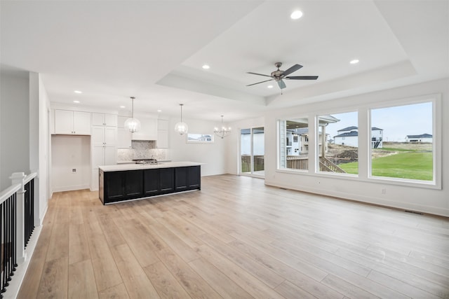 unfurnished living room with ceiling fan with notable chandelier, a raised ceiling, and light wood-type flooring