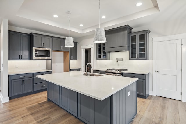 kitchen with stainless steel appliances, a tray ceiling, a kitchen island with sink, sink, and pendant lighting