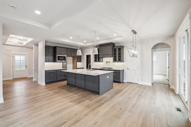 kitchen with light hardwood / wood-style floors, stainless steel microwave, a kitchen island with sink, and hanging light fixtures