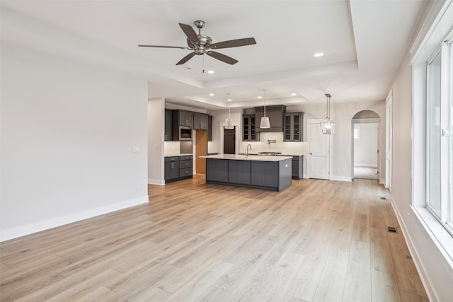 kitchen with ceiling fan, a raised ceiling, pendant lighting, light hardwood / wood-style floors, and a kitchen island with sink