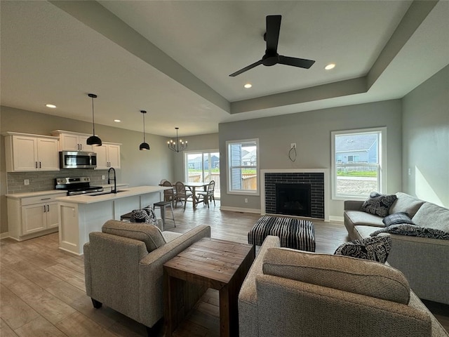 living room with ceiling fan with notable chandelier, sink, a brick fireplace, light wood-type flooring, and a tray ceiling