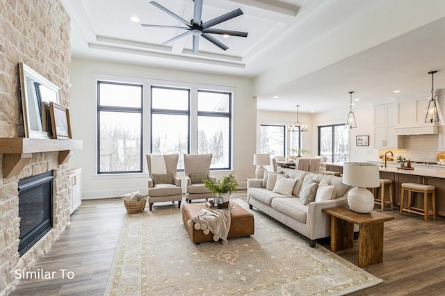 living room with ceiling fan with notable chandelier, a raised ceiling, sink, a fireplace, and wood-type flooring