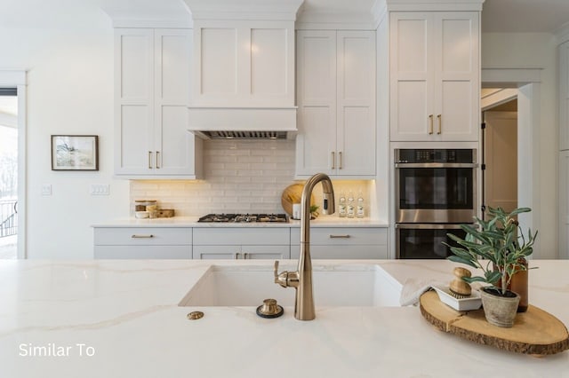 kitchen with white cabinetry, light stone countertops, stainless steel appliances, premium range hood, and decorative backsplash