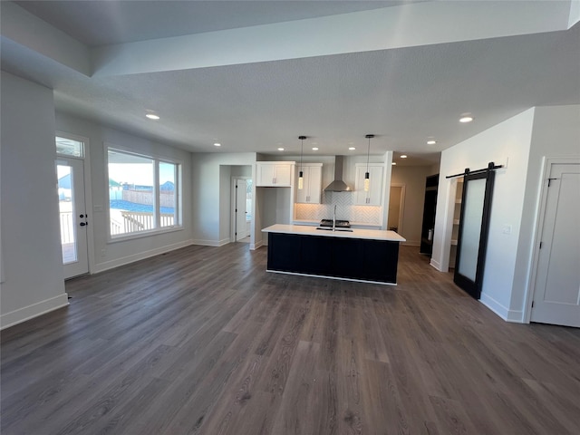 kitchen with a barn door, wall chimney range hood, white cabinetry, hanging light fixtures, and a kitchen island with sink