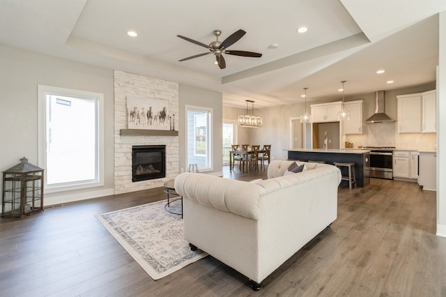 living room featuring dark hardwood / wood-style flooring, ceiling fan, a stone fireplace, and a tray ceiling