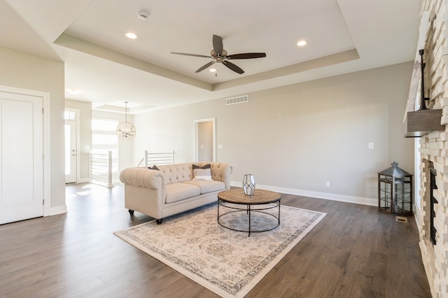 living room with a tray ceiling, a stone fireplace, dark wood-type flooring, and ceiling fan with notable chandelier