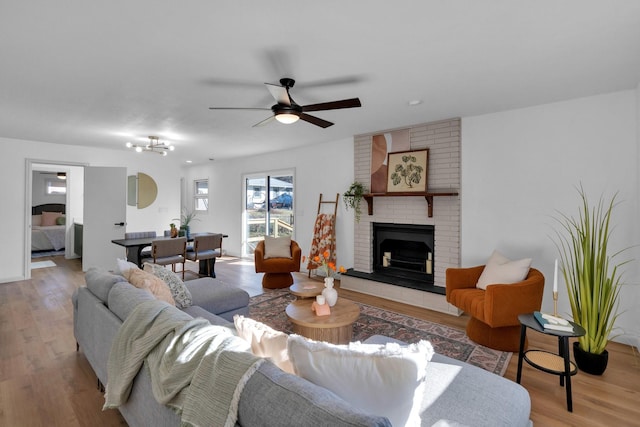 living room with ceiling fan with notable chandelier, light hardwood / wood-style flooring, and a brick fireplace