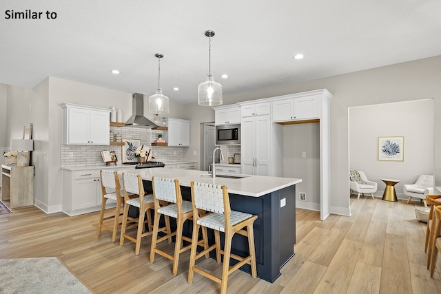 kitchen featuring wall chimney exhaust hood, sink, white cabinets, stainless steel microwave, and an island with sink