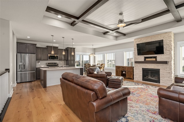 living room featuring coffered ceiling, ceiling fan with notable chandelier, a stone fireplace, light hardwood / wood-style flooring, and beam ceiling