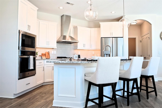 kitchen featuring white cabinets, appliances with stainless steel finishes, an island with sink, and wall chimney range hood