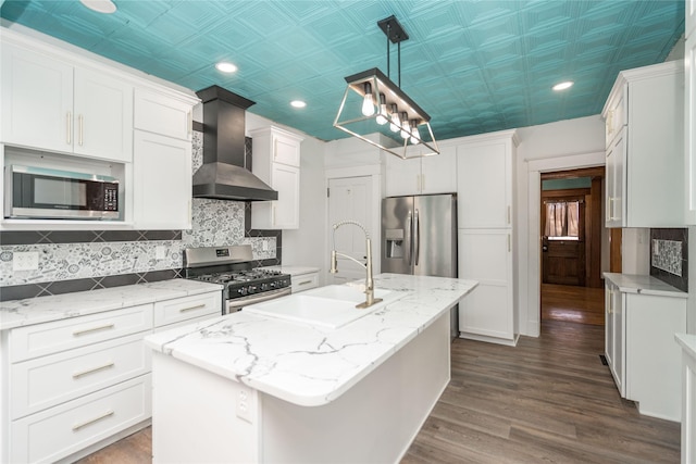 kitchen with appliances with stainless steel finishes, wall chimney exhaust hood, decorative light fixtures, dark hardwood / wood-style floors, and an island with sink