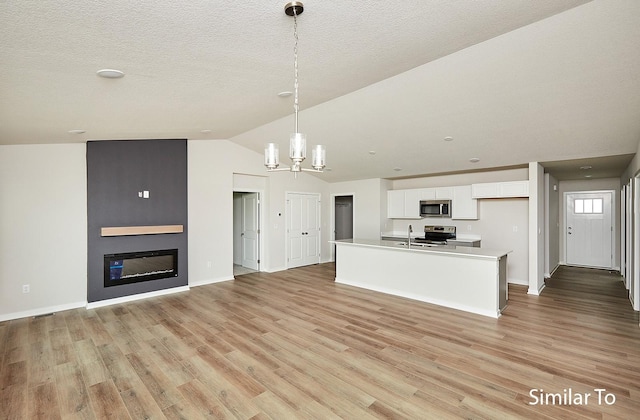 kitchen with appliances with stainless steel finishes, a large fireplace, an island with sink, white cabinets, and decorative light fixtures