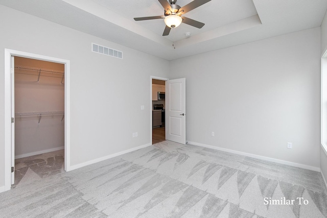 unfurnished bedroom featuring a raised ceiling, a spacious closet, ceiling fan, and light carpet