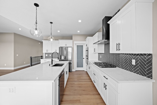 kitchen with wall chimney exhaust hood, stainless steel appliances, a large island with sink, decorative light fixtures, and light hardwood / wood-style flooring