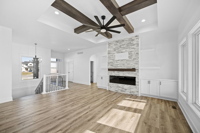 unfurnished living room with ceiling fan with notable chandelier, light hardwood / wood-style floors, a stone fireplace, and beam ceiling