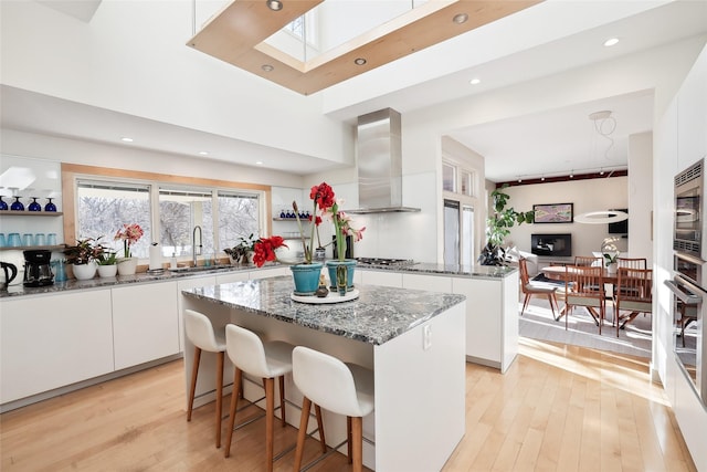 kitchen featuring light wood-style flooring, white cabinets, a center island, wall chimney exhaust hood, and dark stone countertops