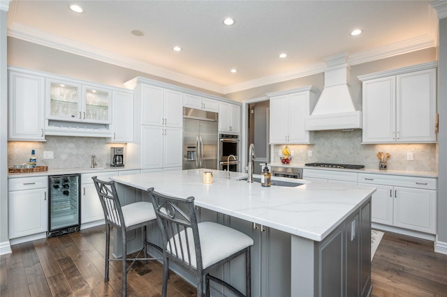 kitchen with white cabinets, appliances with stainless steel finishes, premium range hood, and an island with sink