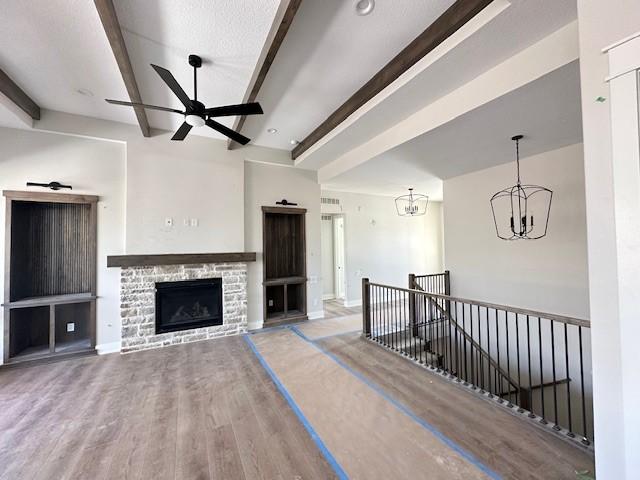 unfurnished living room featuring ceiling fan with notable chandelier, a stone fireplace, wood finished floors, and beam ceiling