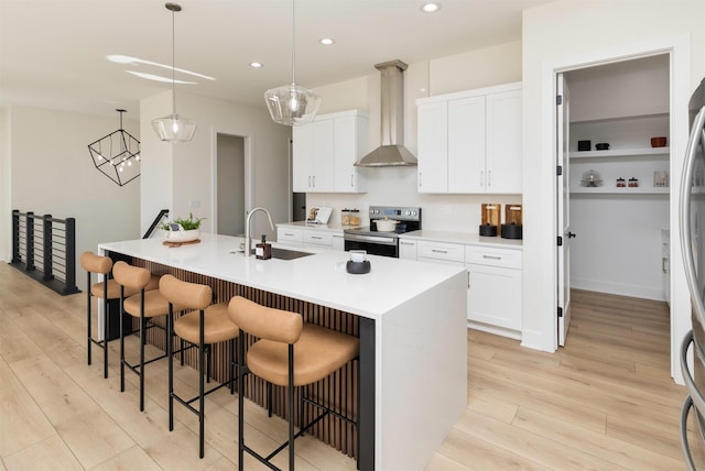 kitchen with sink, stainless steel electric range oven, hanging light fixtures, an island with sink, and wall chimney range hood