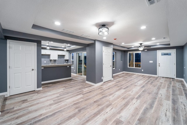 unfurnished living room with a textured ceiling, light wood-type flooring, a raised ceiling, and ceiling fan