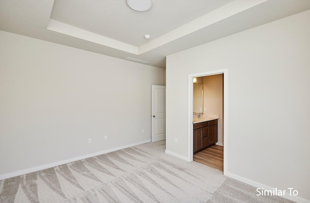 unfurnished bedroom featuring connected bathroom, light carpet, a sink, baseboards, and a raised ceiling