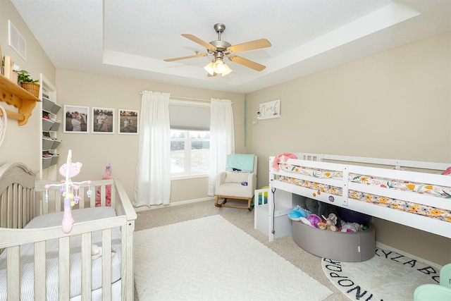 carpeted bedroom with a crib, a tray ceiling, and ceiling fan