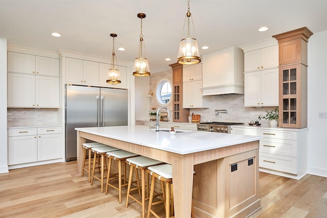 kitchen with built in fridge, a sink, light wood-style floors, a large island with sink, and custom range hood