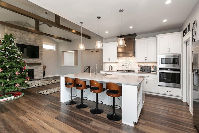 kitchen featuring a kitchen island with sink, premium range hood, hanging light fixtures, appliances with stainless steel finishes, and beamed ceiling