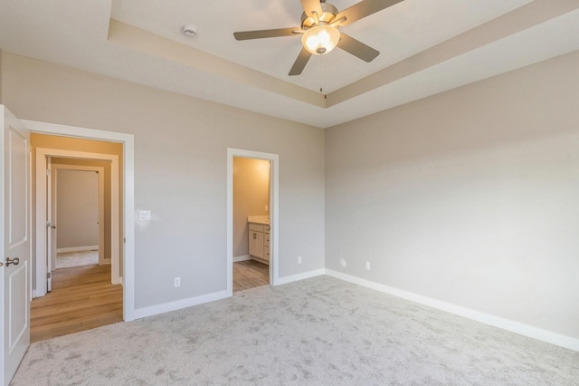 unfurnished bedroom with a tray ceiling, ensuite bath, ceiling fan, and light carpet