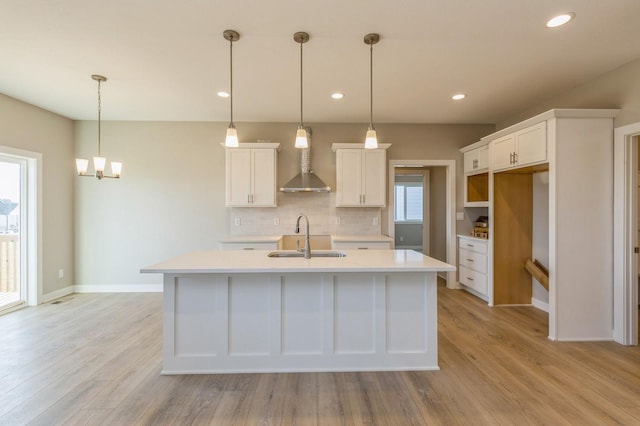 kitchen with sink, wall chimney exhaust hood, an island with sink, decorative light fixtures, and white cabinets