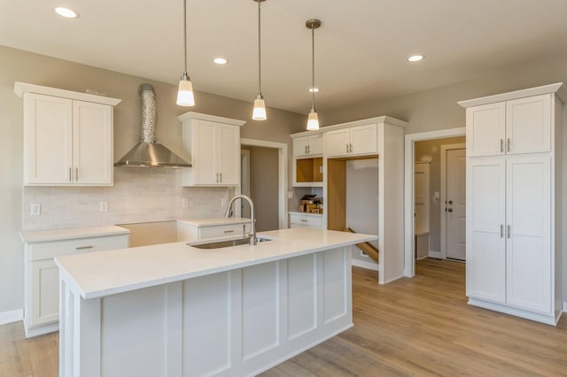 kitchen with sink, wall chimney range hood, white cabinets, hanging light fixtures, and an island with sink