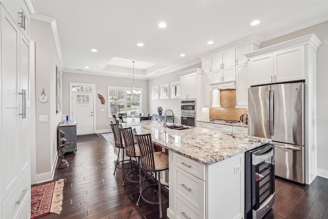 kitchen with wine cooler, stainless steel appliances, a raised ceiling, a kitchen island with sink, and white cabinets