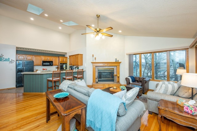 living room with a skylight, ceiling fan, high vaulted ceiling, light hardwood / wood-style floors, and a tiled fireplace