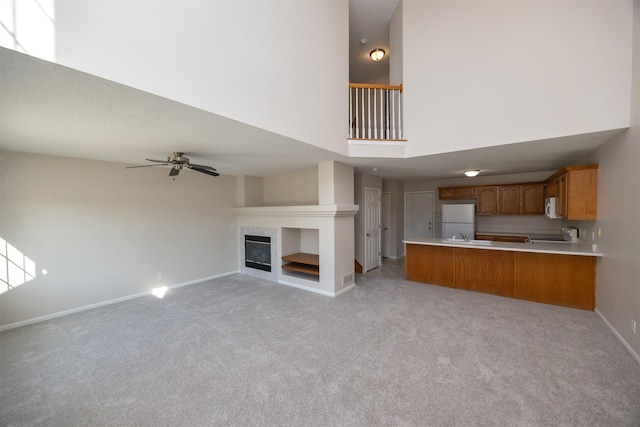 unfurnished living room with a towering ceiling, a glass covered fireplace, light carpet, ceiling fan, and baseboards