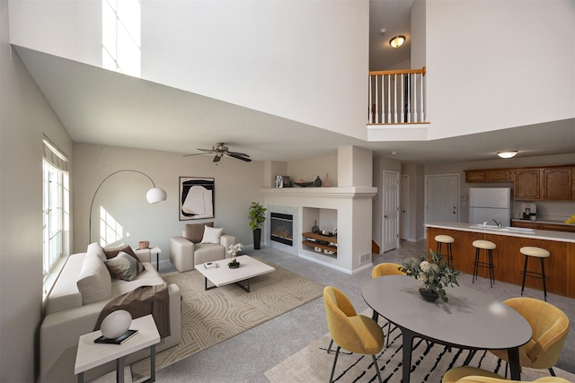 living room featuring baseboards, light colored carpet, ceiling fan, a high ceiling, and a fireplace